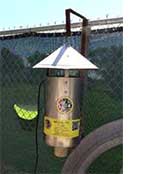 Automatic mosquito trap hanging on a metal stand with a green leaf in the foreground and a chain-link fence in the background.