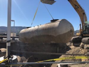 Large cylindrical tank being lifted by an excavator at a construction site, with caution tape around the area.