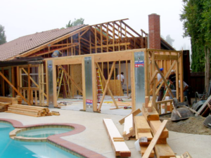 Framed construction work on a house near a pool. Two ladders, wooden beams, and construction tools are visible.