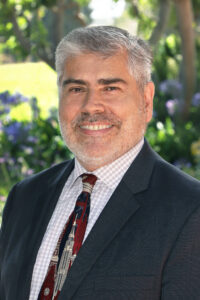 Portrait of a smiling man with grey hair, wearing a dark suit, patterned tie, and checked shirt, with green foliage and flowers in the background