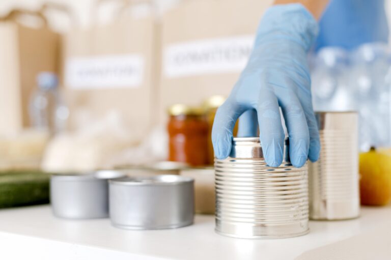 A hand wearing a blue glove holding a tin can, with other cans, food, and donation bags in the background.