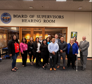 A group of individuals standing together in front of the entrance to the Board of Supervisors Hearing Room, located in Ventura County, California. The sign above the entrance displays the name of the room.