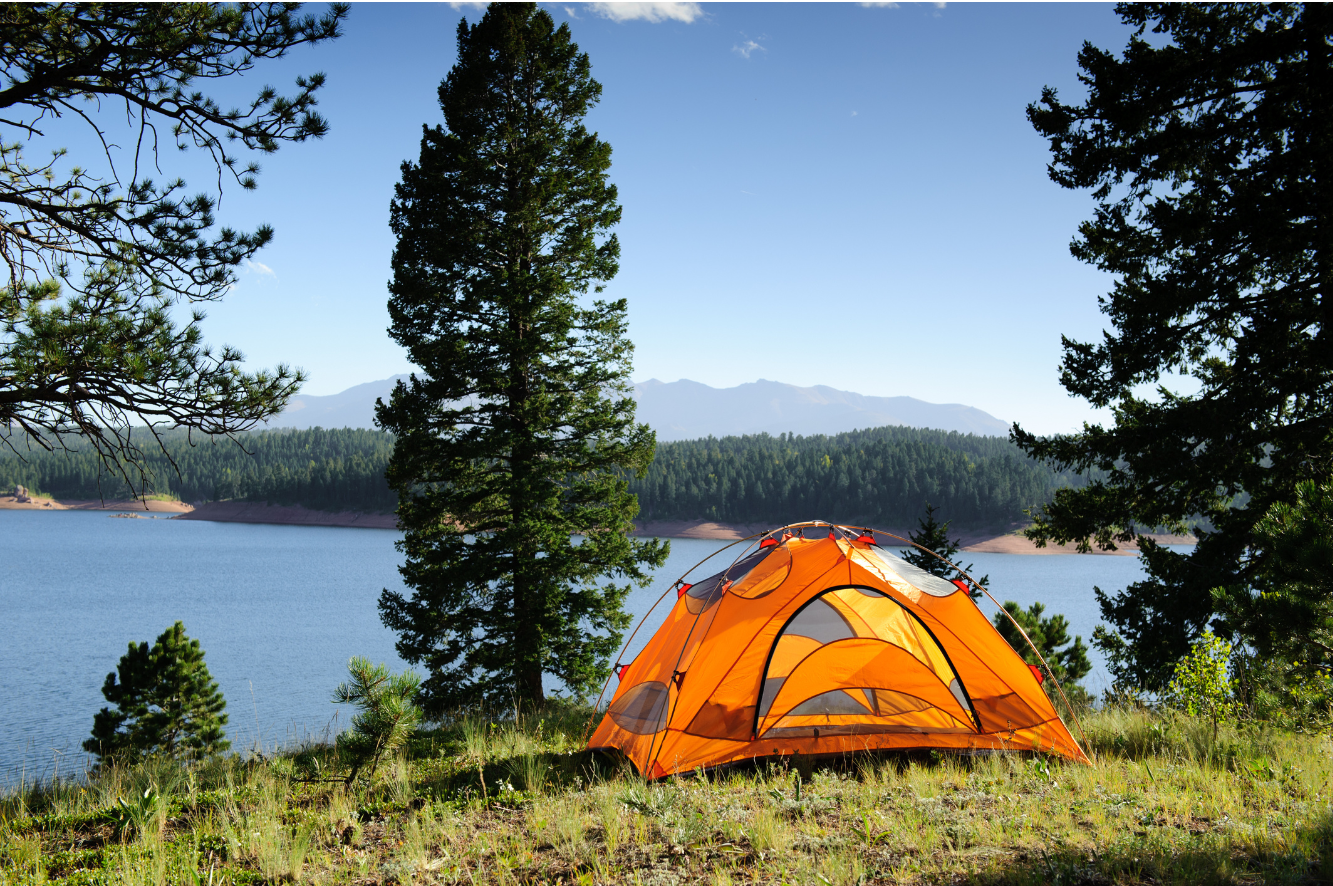 Orange tent pitched by a lakeside in a forested area with mountains in the background