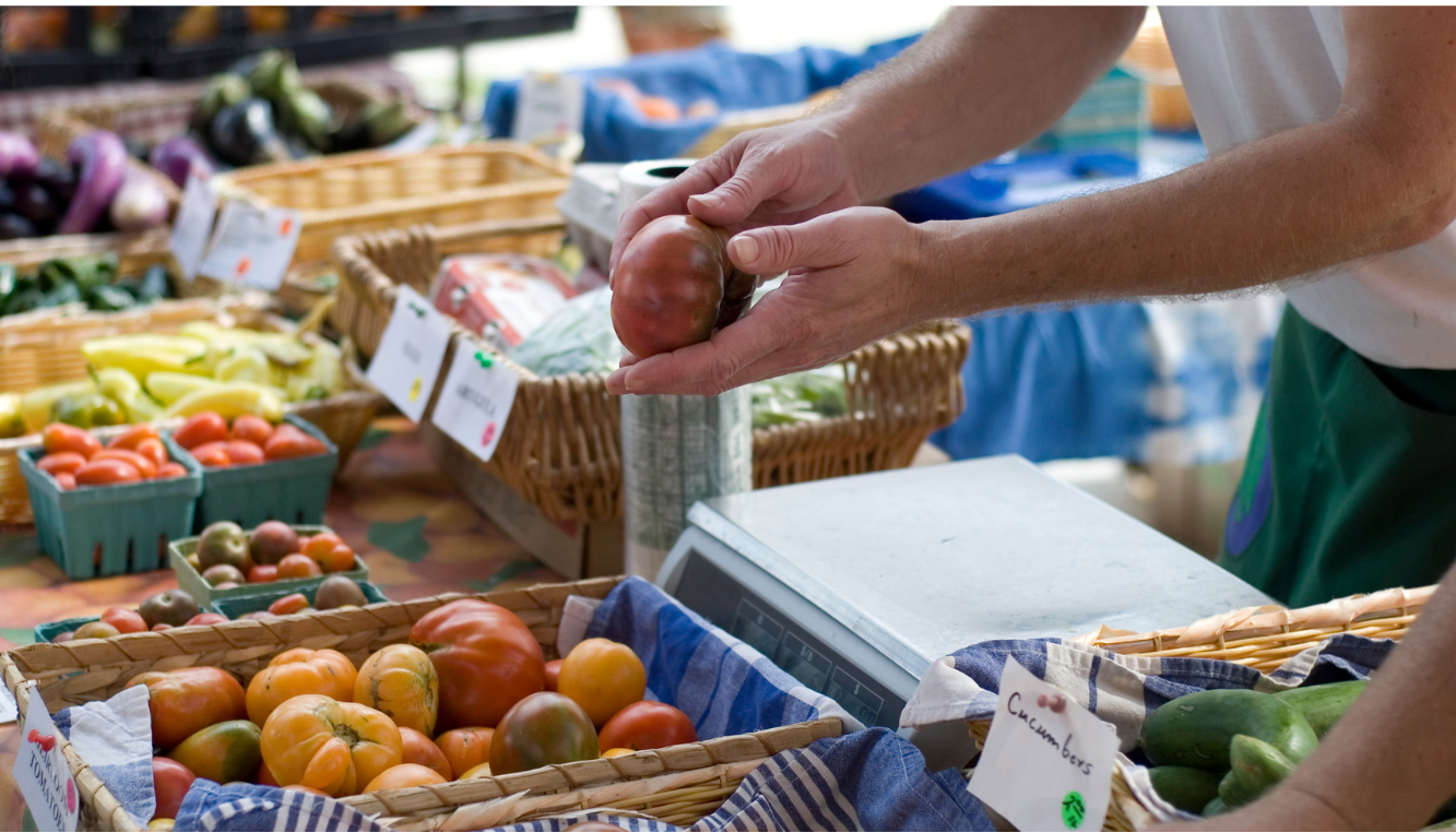 Hands holding a tomato at a farmers market stall displaying various fresh vegetables.