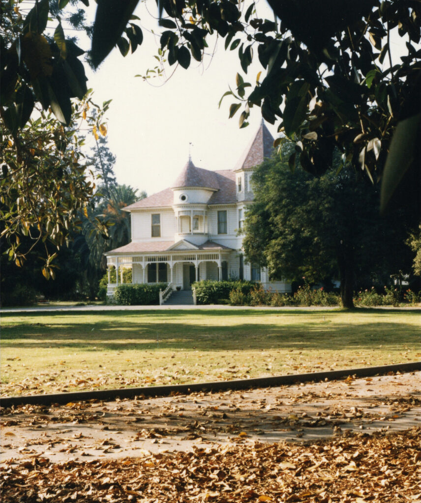 View of front yard with grass and mature trees framing the Victorian Camarillo House, circa 1980s.