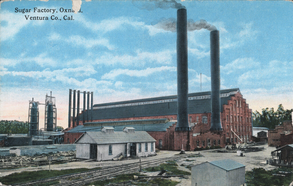 Oblique view of the former Oxnard Sugar Beet Factory, featuring smokestacks and surrounding buildings.