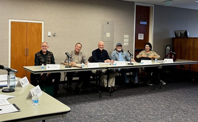 Five people seated at a conference table with microphones and nameplates, engaging in a local government meeting.